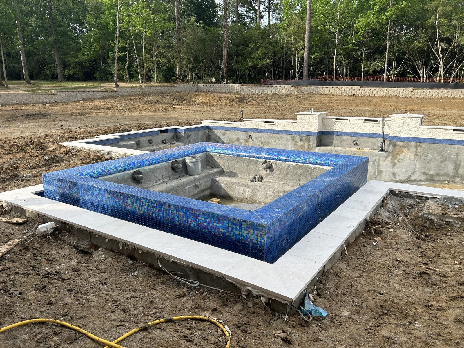 Raised spa wrapped in iridescent blue glass tile beside the unfinished pool shell and masonry back wall